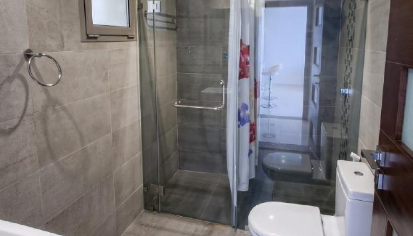 Modern bathroom with gray tiles, glass shower enclosure, curtain, toilet, small window, towel ring, and sink.