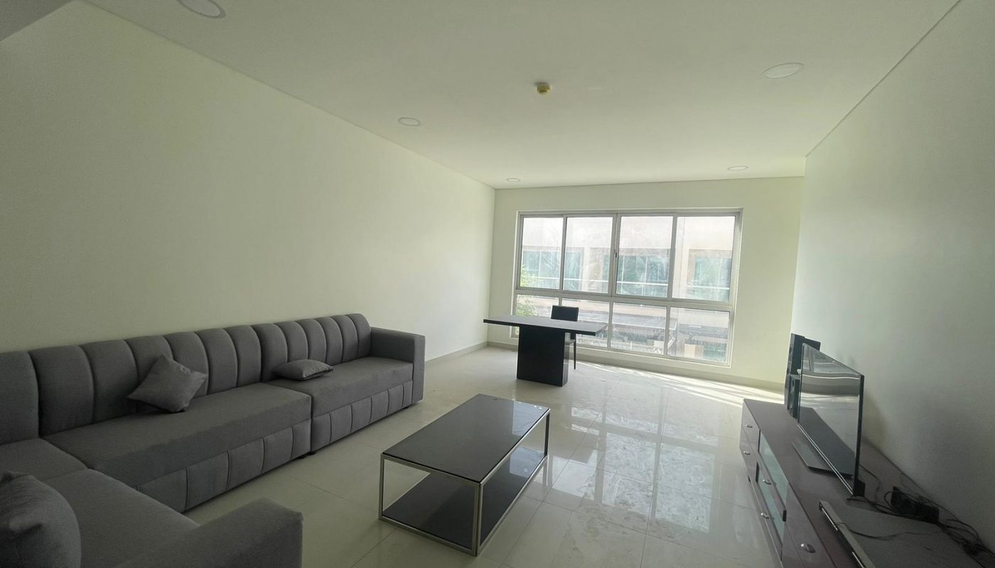 A modern, minimalist living room with a gray sectional sofa, glass coffee table, TV on a stand, and a desk by a large window letting in natural light.