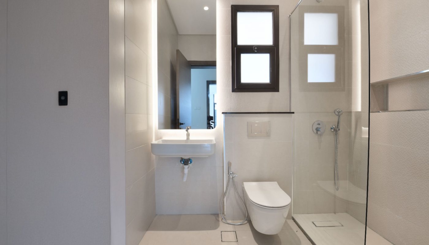 Modern bathroom with a wall-mounted sink, toilet, bidet spray, glass-enclosed shower, and two frosted windows. Neutral color scheme with minimalistic design.