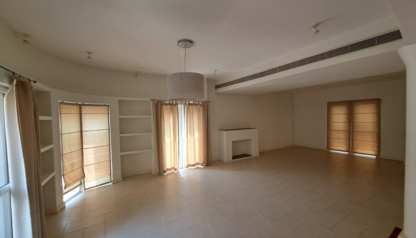 Empty living room with tiled floors, beige walls, a hanging light fixture, shelves, covered windows, and a small fireplace. Natural light enters through the window coverings.