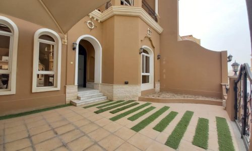 A luxury multi-story villa for rent in Janabiya, featuring arched windows, a tiled entrance, and a patio with evenly spaced grass strips. The building's beige exterior with white trim adds to its elegant charm.