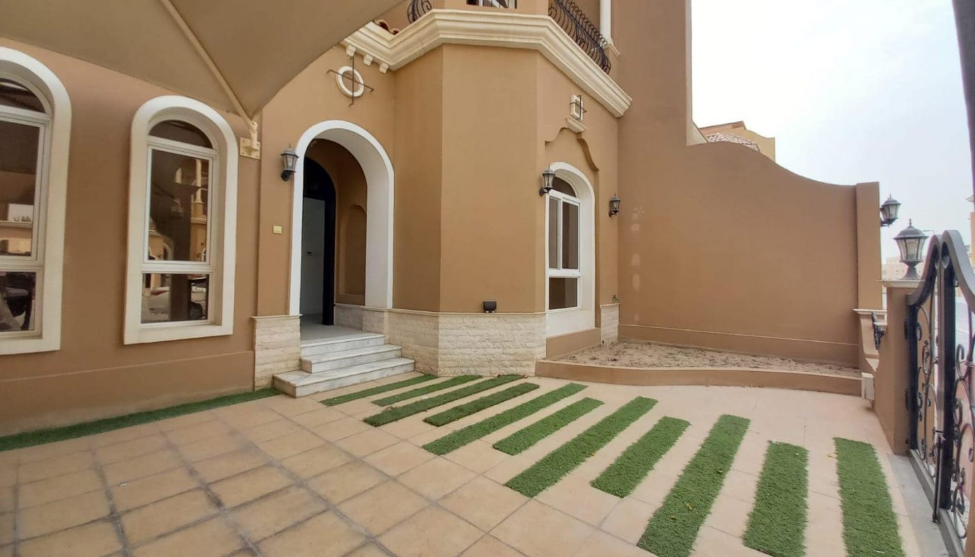A luxury multi-story villa for rent in Janabiya, featuring arched windows, a tiled entrance, and a patio with evenly spaced grass strips. The building's beige exterior with white trim adds to its elegant charm.