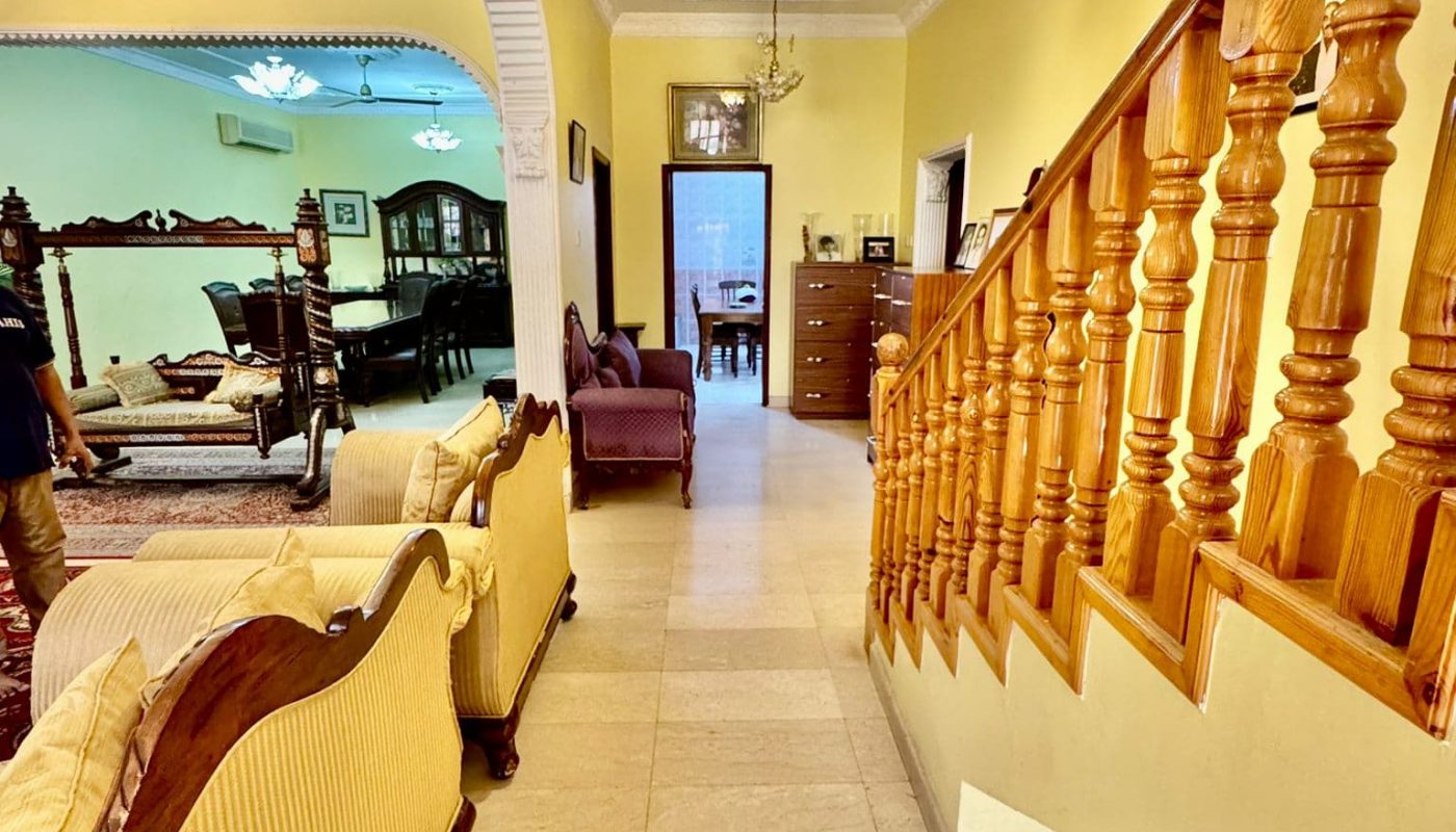 A hallway with tiled flooring, yellow walls, a wooden staircase on the right, and vintage-style sofas leading to a green-painted living room and a lit chandelier overhead.