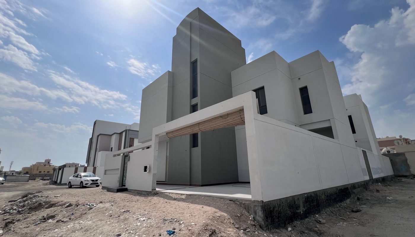 Modern two-story white house with a flat roof, large windows, and a gated driveway under a bright, partly cloudy sky. A white car is parked nearby on a dirt lot.