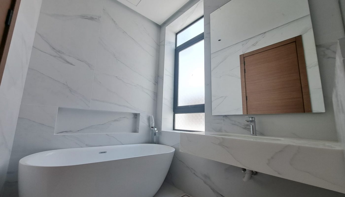 Modern bathroom with a white freestanding bathtub, large wall mirror, marble walls and floors, and a window next to a wooden door.