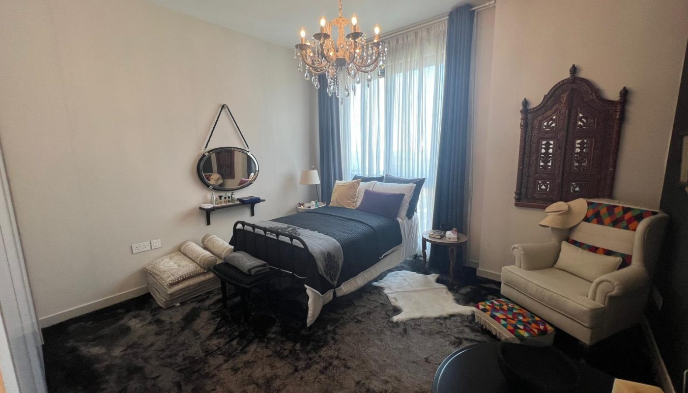 A neatly arranged bedroom with a single bed, chandelier, armchair, wall mirror, ornate wooden wall decor, and a window with sheer curtains letting in natural light.