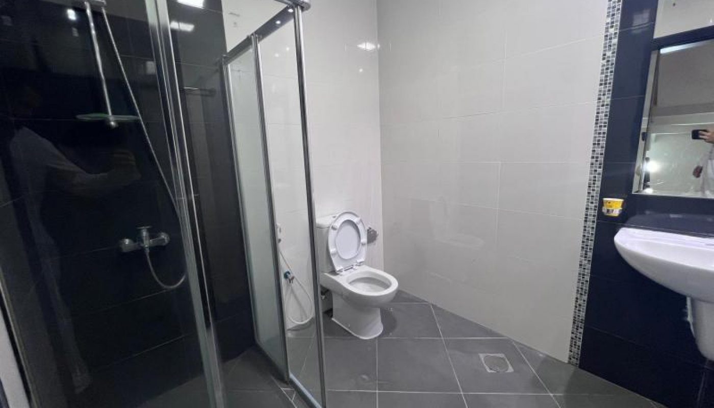A clean modern bathroom with a glass shower enclosure, a toilet with the lid up, a wall-mounted sink, and large gray floor tiles.