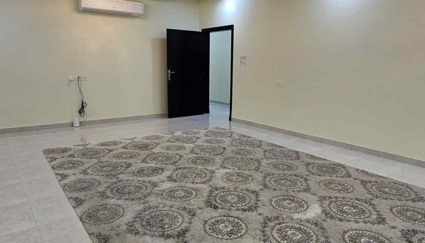 A spacious, empty room with tiled floors, a patterned area rug, two chandeliers, cream-colored walls, a ceiling fan, an air conditioner, and an open black door.