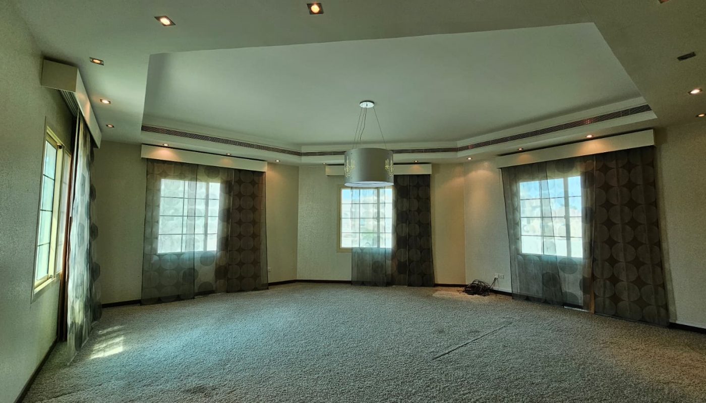 Spacious empty room with carpeted floor, three large windows with patterned curtains, ceiling lights, and a central hanging light fixture.
