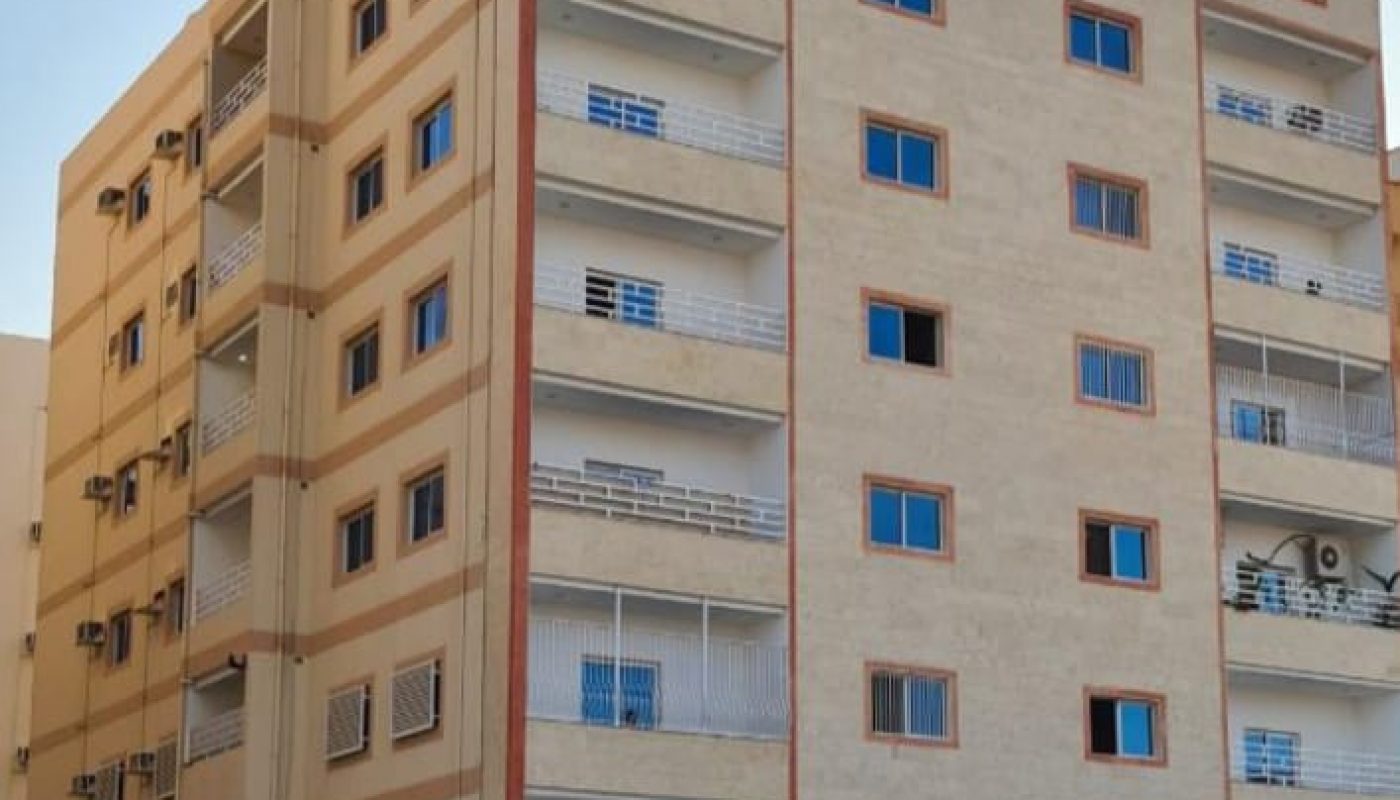 مبنى سكني من ستة طوابق باللون البيج مع شرفات متعددة ونوافذ ذات قضبان حديدية، تم تصويره من زاوية في ضوء النهار.