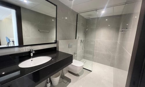 Modern bathroom interior in an Amwaj apartment for rent, featuring a wall-mounted sink, large mirror, toilet, and a glass-enclosed shower area, with gray tiles and ambient lighting.