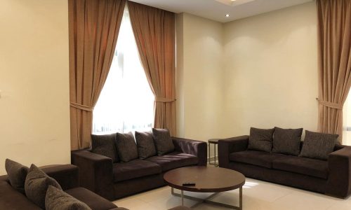 A living room with three brown sofas, a round wooden coffee table, and two windows with beige curtains. The room has tiled floors and ceiling lights.