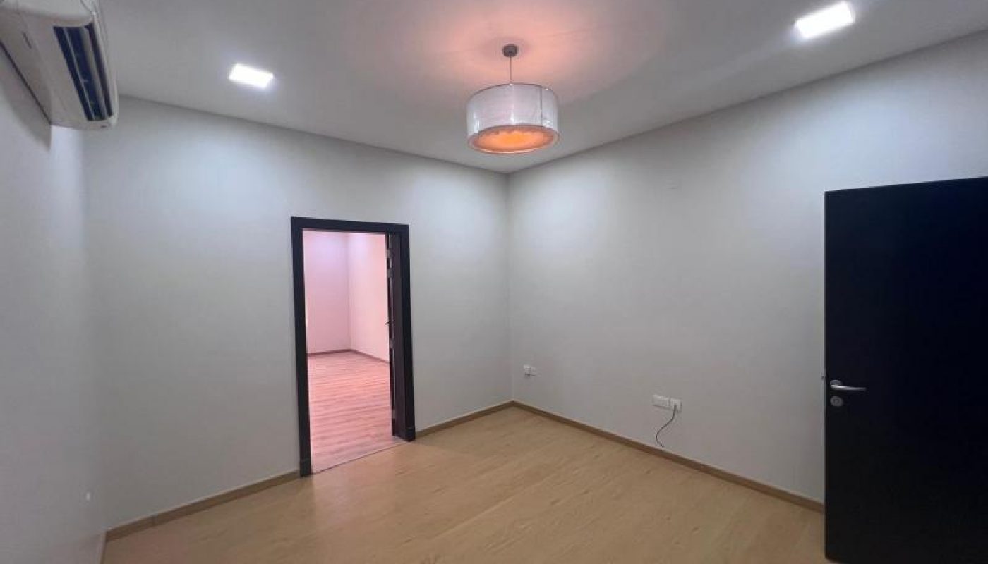 Empty room with light wood flooring, white walls, a ceiling light fixture, an air conditioner, an open black door, and a doorway leading to another room.