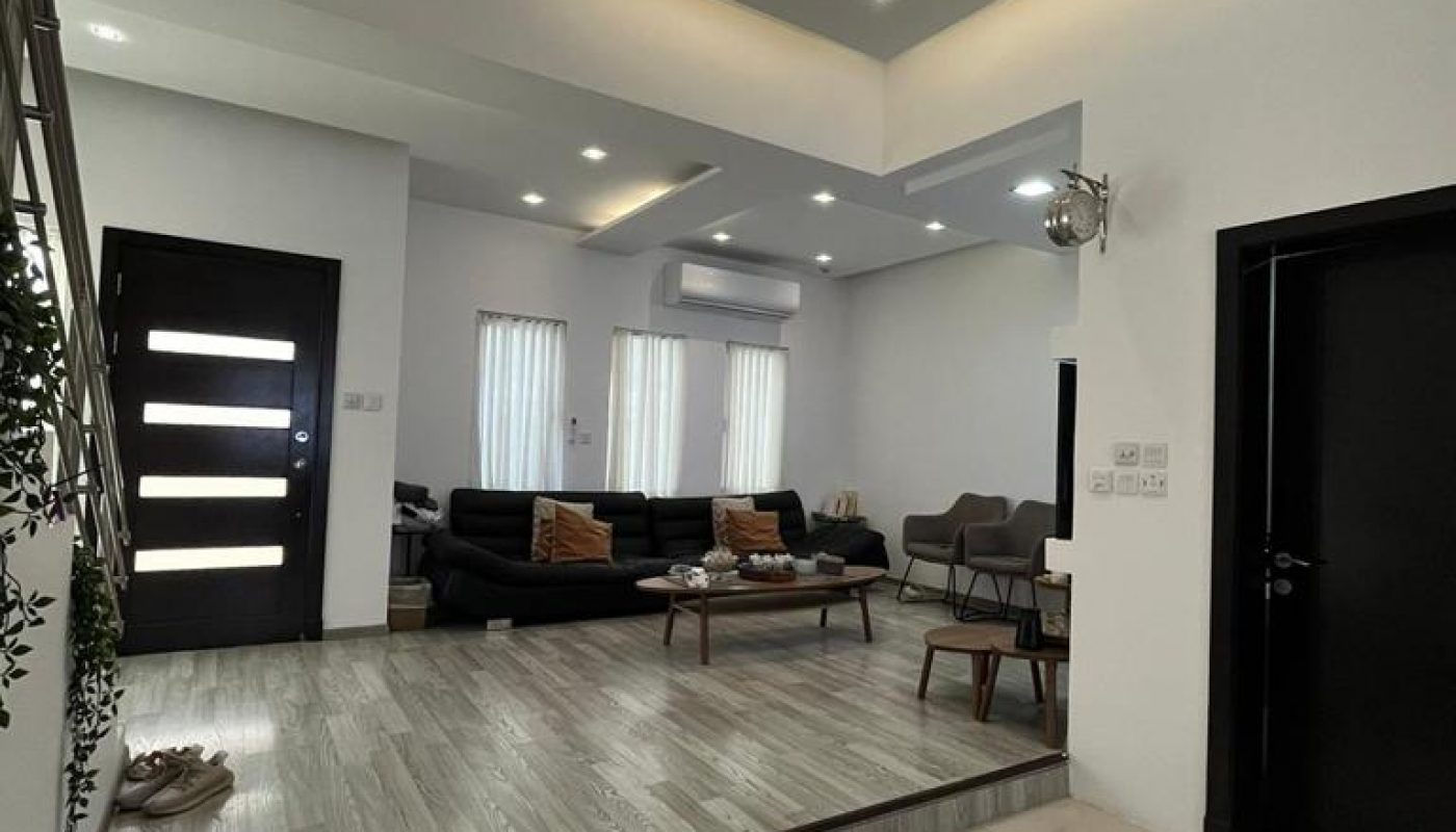 Modern living room with recessed lighting, black sofas, accent chairs, wood flooring, and white tiled entryway. Large windows let in natural light. Door and staircase visible on the left.