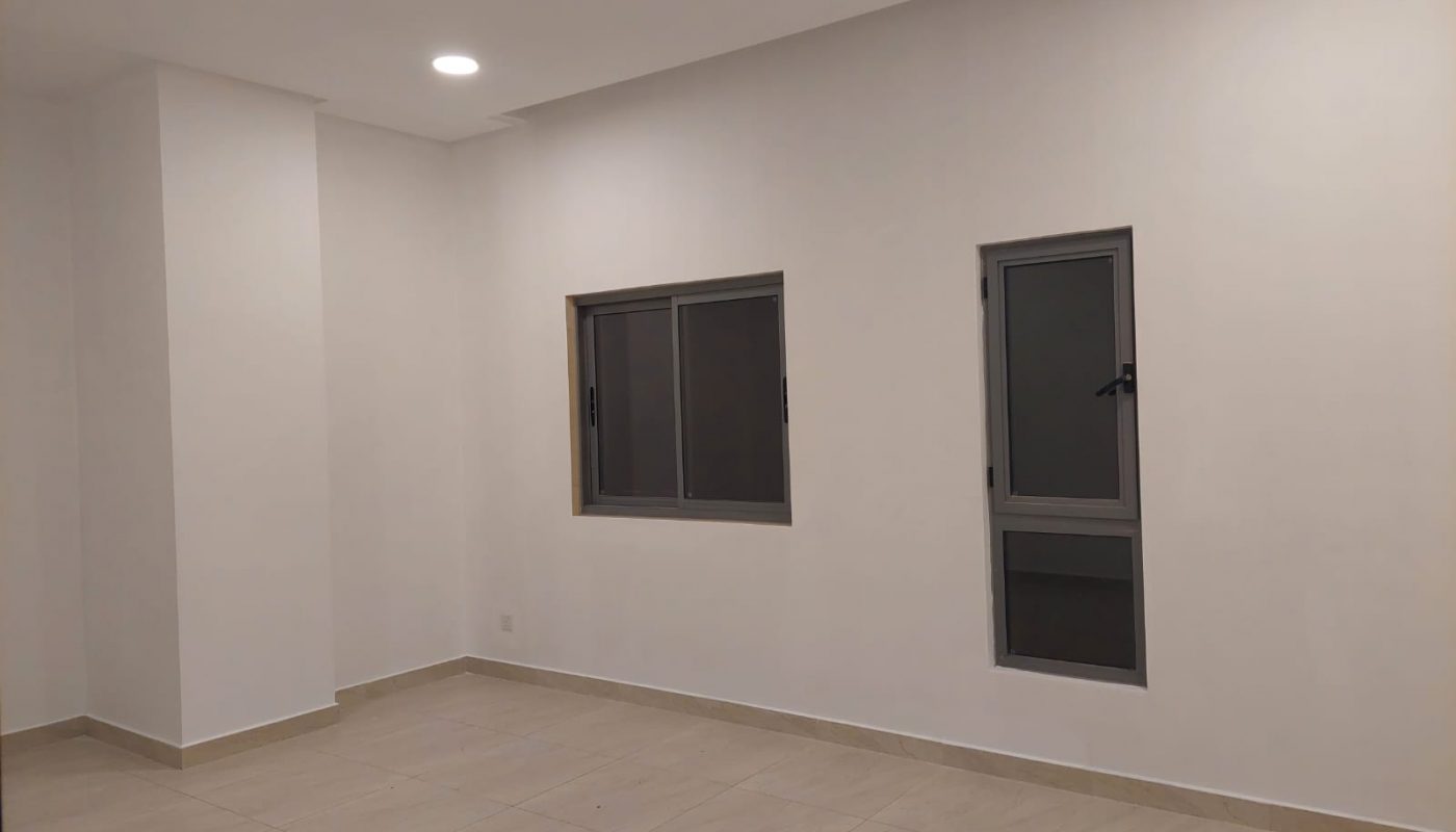 Empty room with beige tiled floor, white walls, a small window and a narrow vertical window. Ceiling light is on.