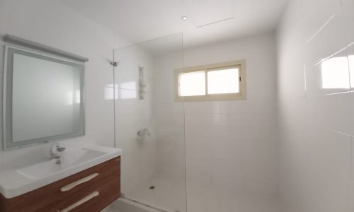 A minimalist bathroom features white walls, a mirror above a wooden cabinet with a sink, and a glass shower enclosure with a window providing natural light.