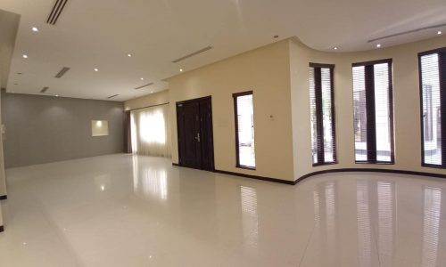 A spacious, unfurnished room with white tiled floor, beige walls, a central dark wooden door, large windows with blinds, and ceiling lights.