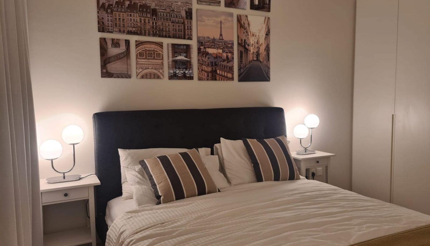 A tidy bedroom in a flat for sale, featuring a double bed, striped cushions, bedside tables with lamps, and a wall decorated with framed photos of cityscapes. Ideal for anyone seeking comfort and style in the heart of Diyar Al-Muharraq.