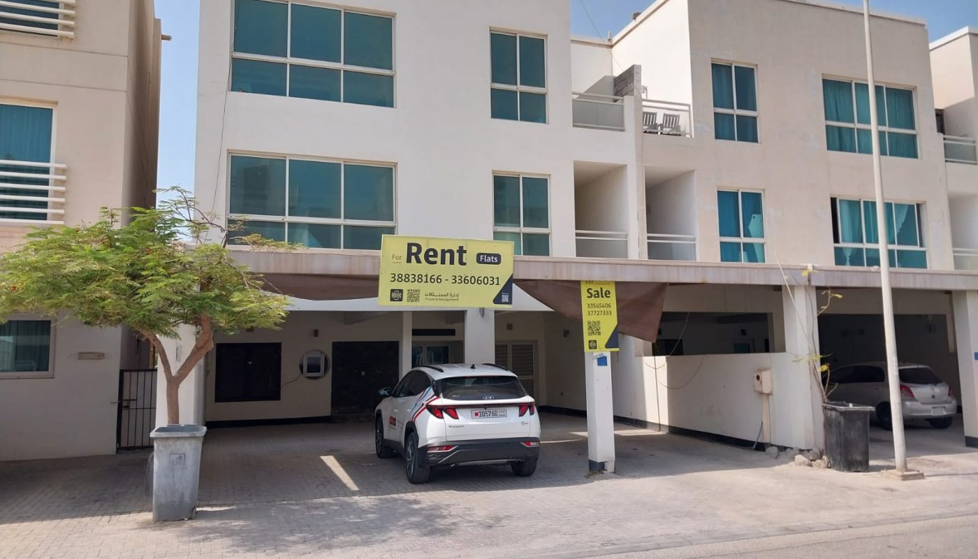 A white modern townhouse with large windows has "For Rent" and "For Sale" signs displayed. Two cars are parked in the driveway under a carport.