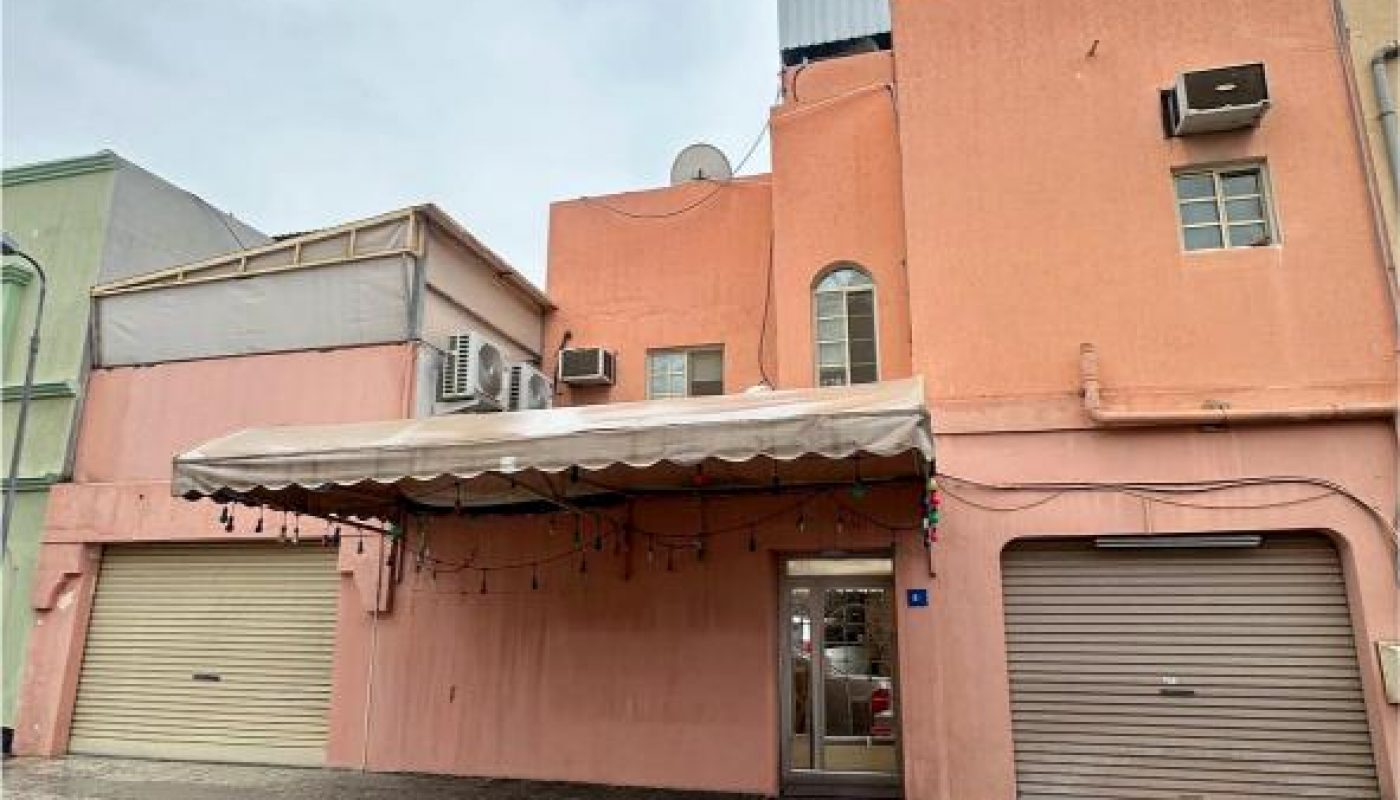 A two-story peach-colored building with closed garage doors, a small entrance, outdoor air conditioning units, and string lights under an awning on a wet street.