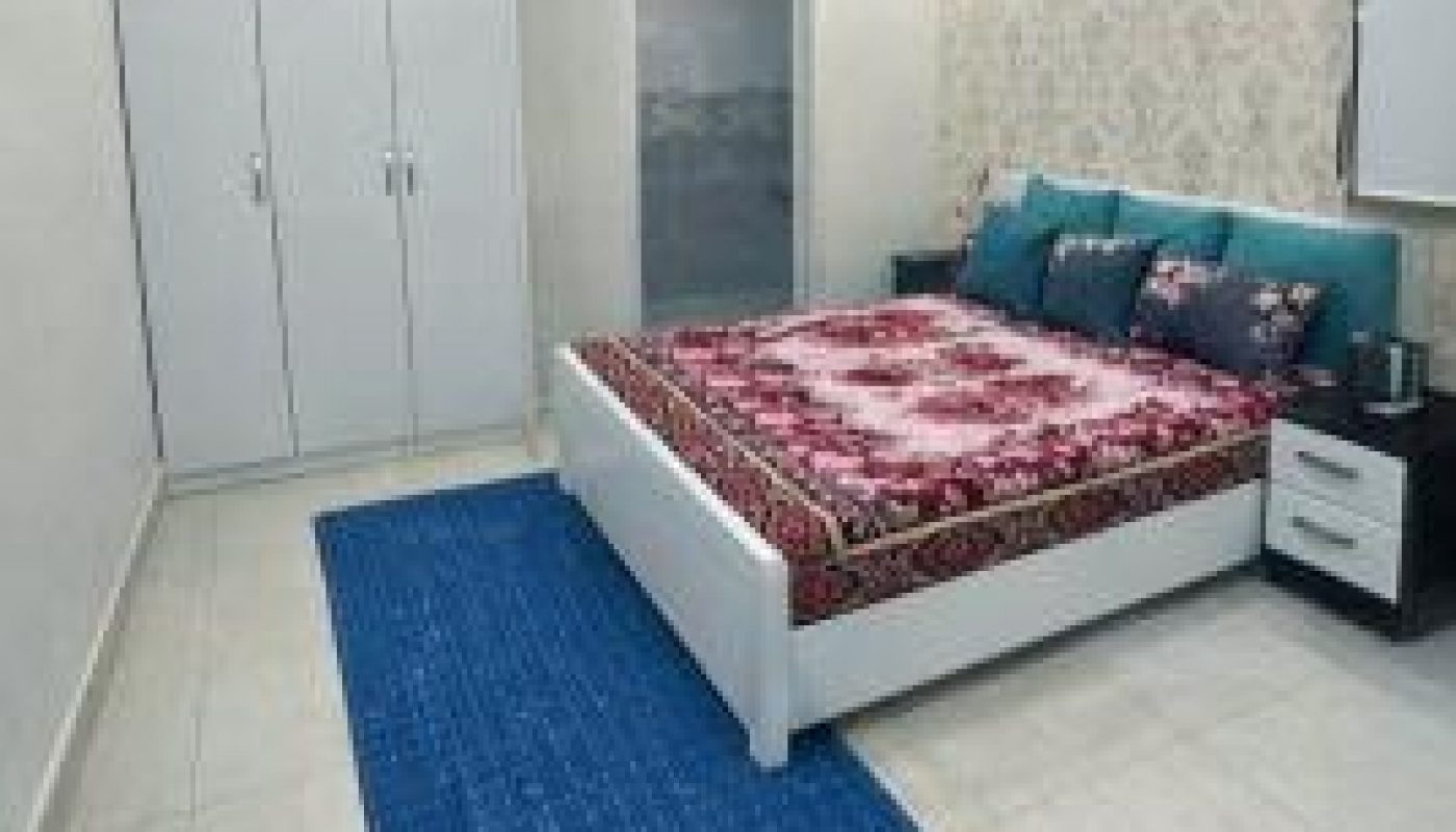A modern bedroom with a white wardrobe, a double bed with a red patterned bedspread, teal pillows, two nightstands, and a blue rug on a tiled floor.