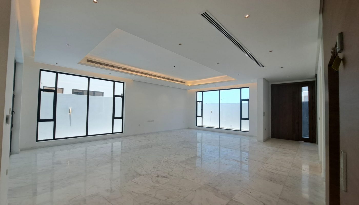 Spacious, empty room with marble floors, large windows, white walls, recessed lights, and a wooden door. Natural light fills the modern, minimalist interior.