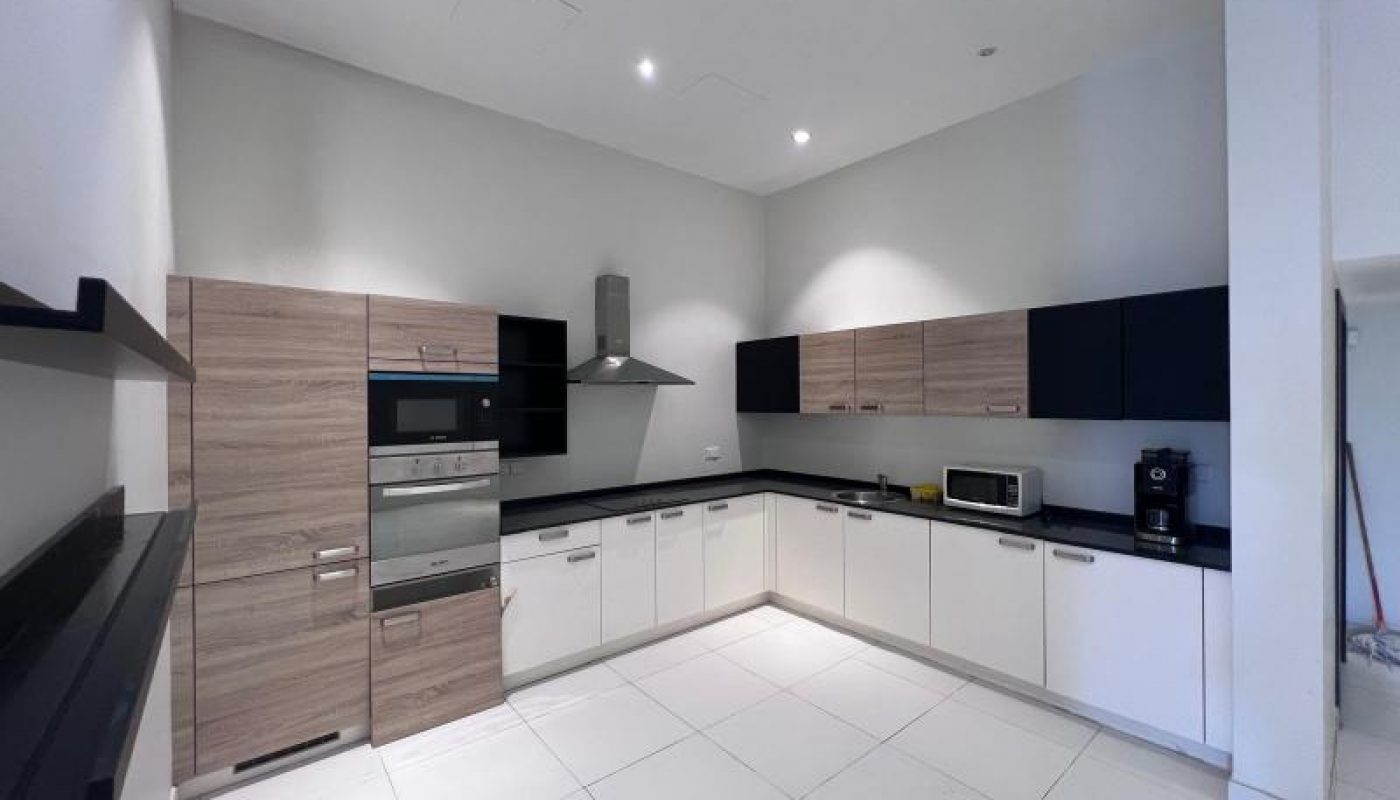 Modern kitchen with light wood and white cabinets, black countertops, built-in oven, microwave, coffee maker, and stainless steel range hood under bright ceiling lights.