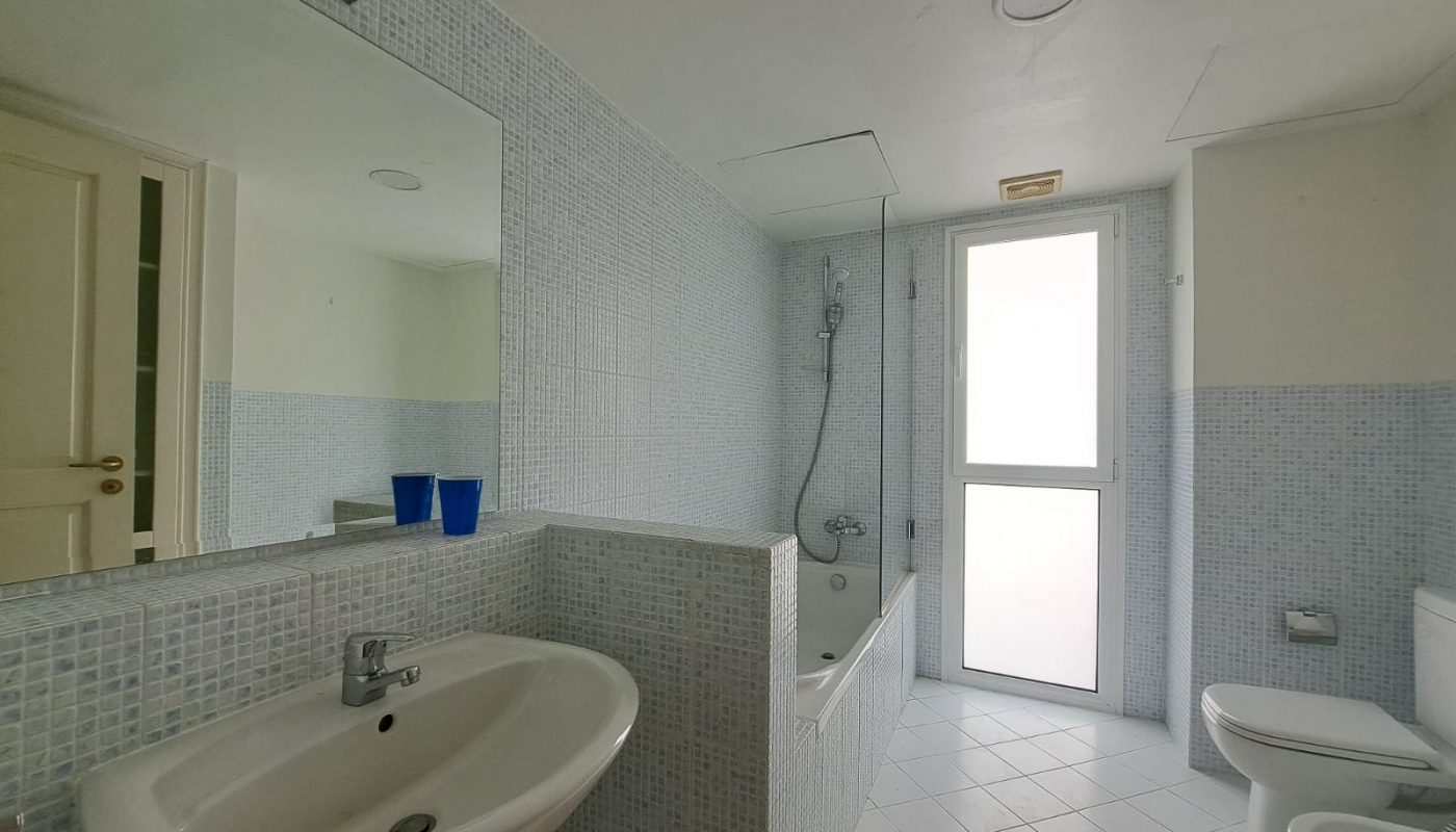 A bathroom with a sink, mirror, two blue cups, bathtub with shower, toilet, bidet, and a frosted glass door letting in natural light.