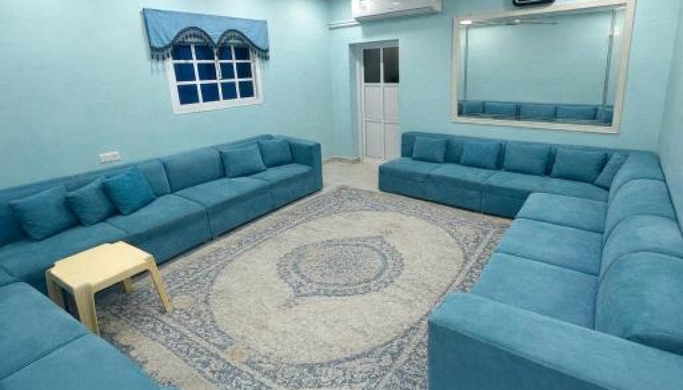 A living room with blue sectional sofas along the walls, a patterned rug in the center, a small beige table, a large mirror, a wall clock, and a window with blue curtains.