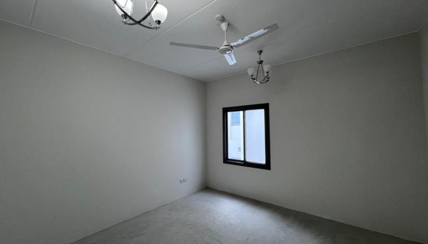 Empty room with plain white walls, a ceiling fan, two chandeliers, a single window, and a bare floor.