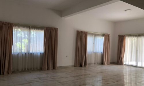 An empty room with white walls, tiled floor, and large windows with beige curtains, allowing natural light to come in.