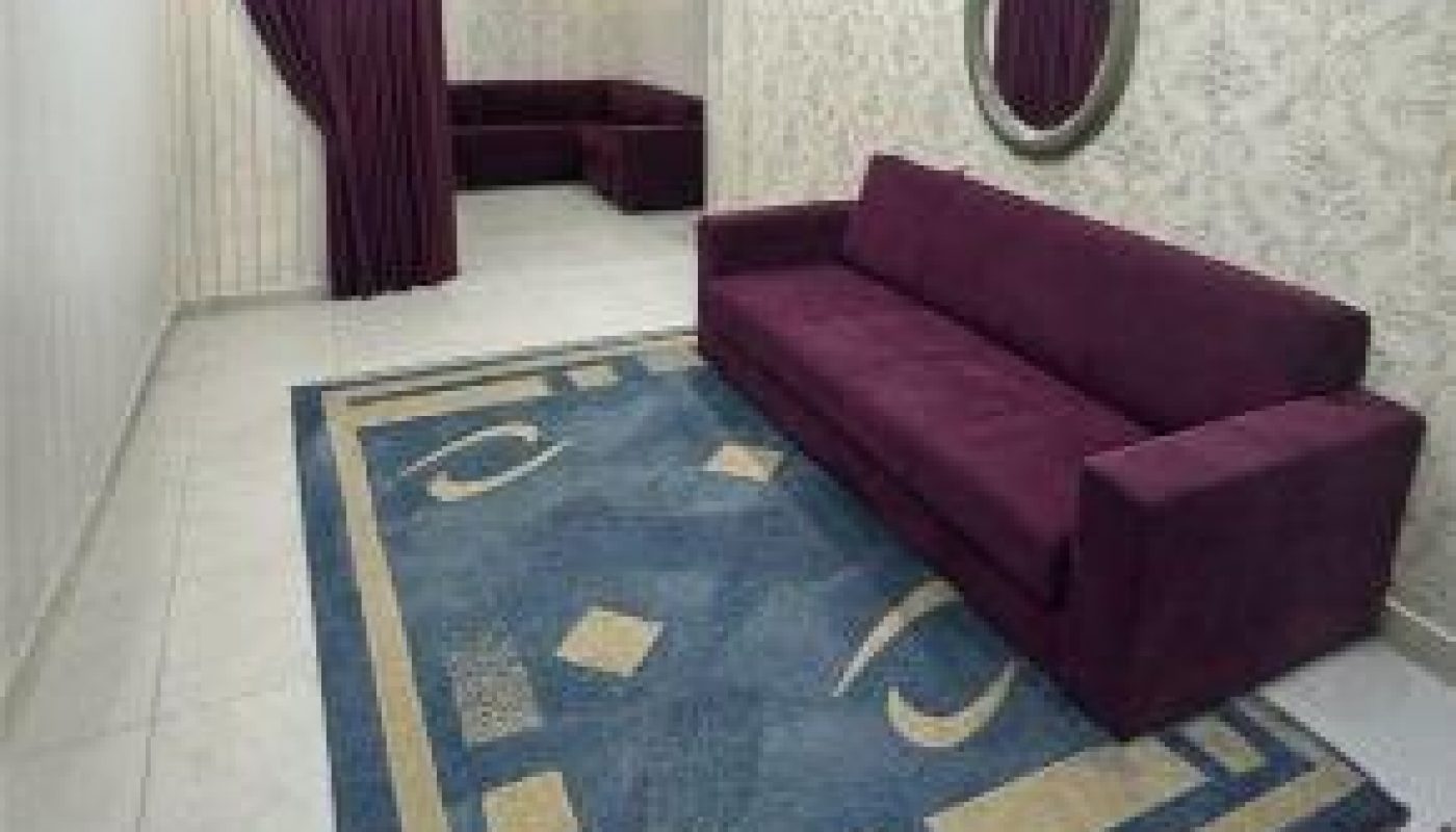 A narrow living room with a maroon sofa, blue patterned rug, wall mirror, and maroon curtains leading to a small alcove with seating.