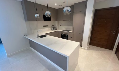 Modern kitchen interior in a luxurious apartment with gray cabinets, a white island, built-in appliances, and pendant lights, featuring a tiled floor and a wooden door.