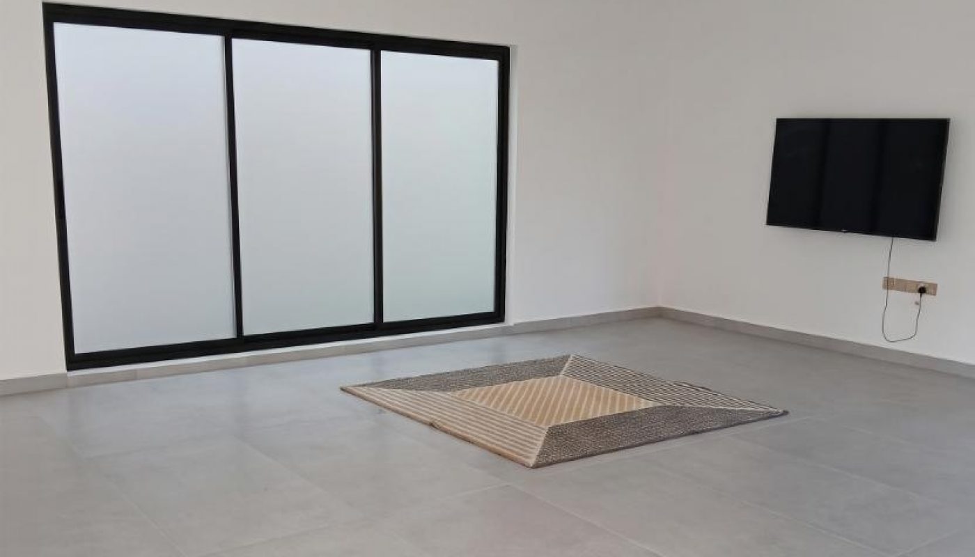 Minimalist room with large frosted window, mounted TV, and a small rug on a light gray tiled floor. Walls are white, and the space is mostly empty.