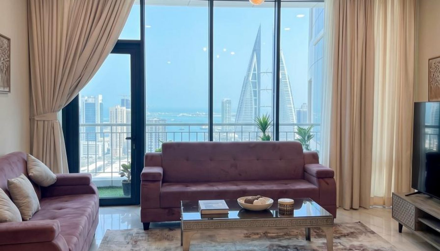 Modern living room with two maroon sofas, a glass coffee table, large windows with city and sea view, light beige curtains, and a TV on a wooden stand.