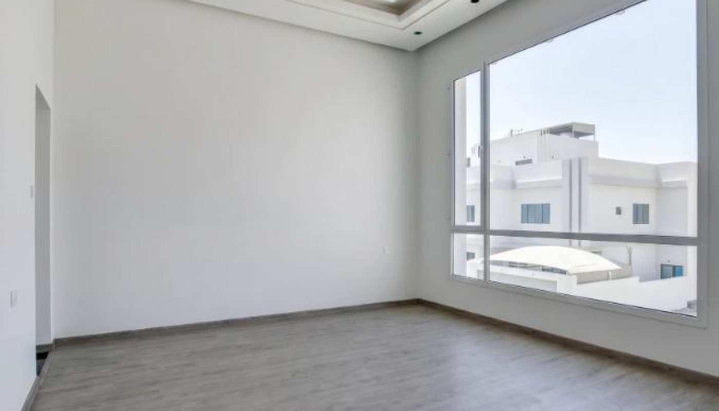 Empty modern room with light gray wood flooring, large window, white walls, and a geometric ceiling light fixture.