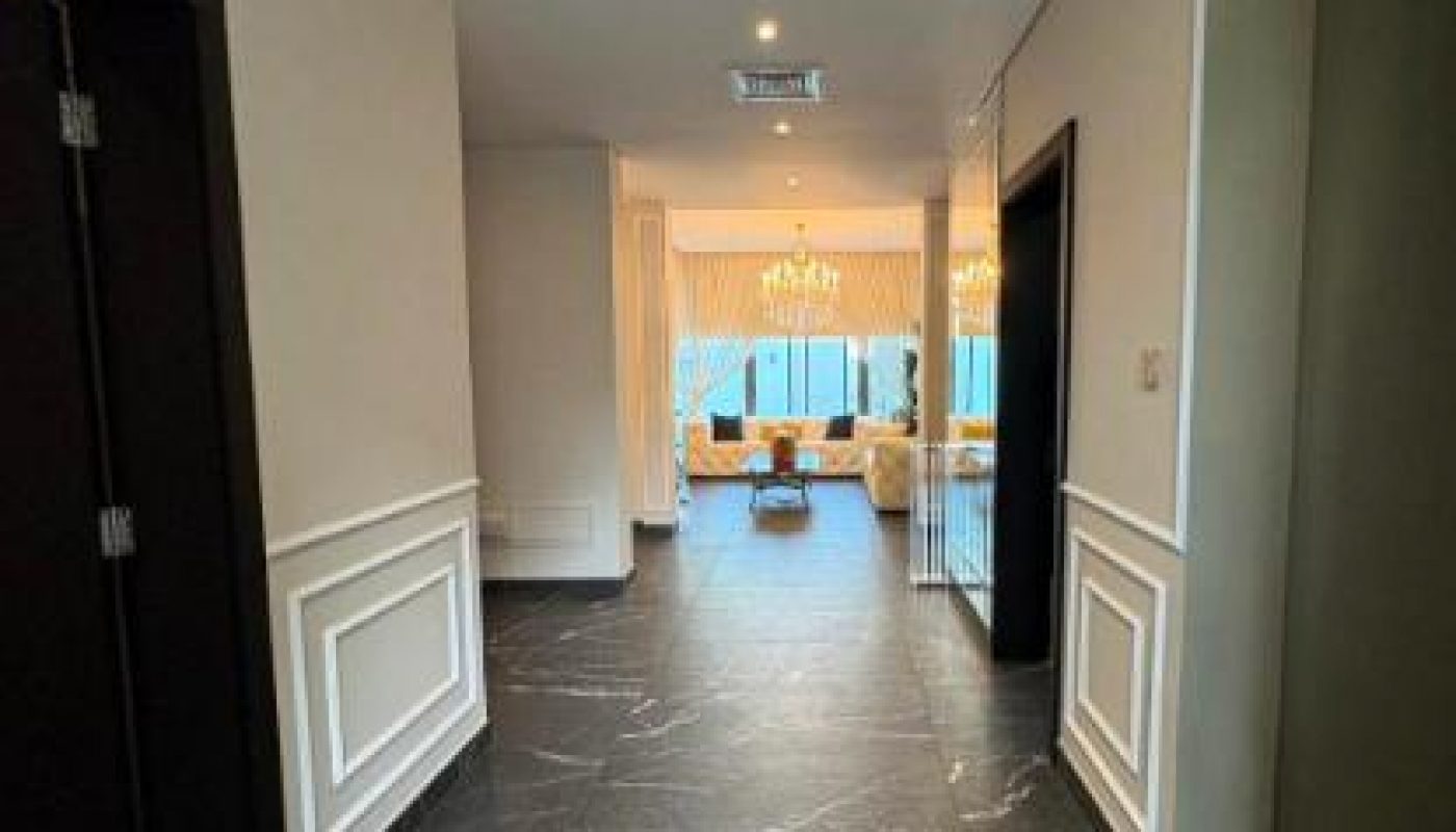 A hallway with dark marble floors and white walls leads to a bright living room with large windows, curtains, and a chandelier.
