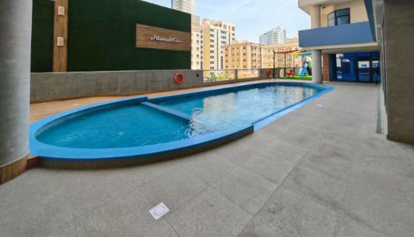 Outdoor swimming pool beside a modern apartment building, surrounded by concrete flooring, with city buildings visible in the background under a clear sky.