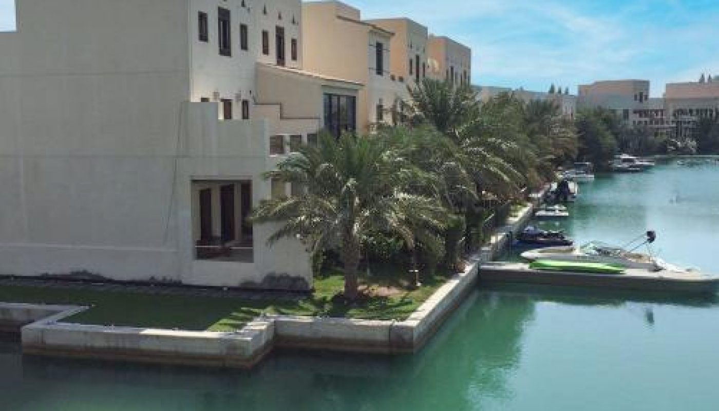 Waterfront villas with palm trees line a canal under a clear blue sky, viewed from behind a decorative balcony railing.