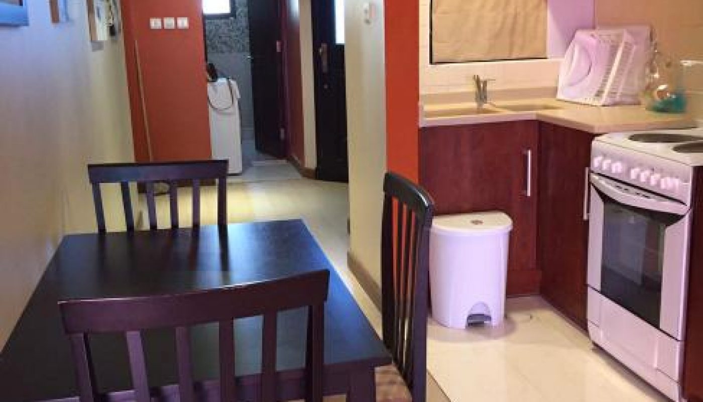 A small kitchen with wooden cabinets, a stove, a sink, a trash bin, and a dining table with three chairs; a washing machine is visible through the doorway.