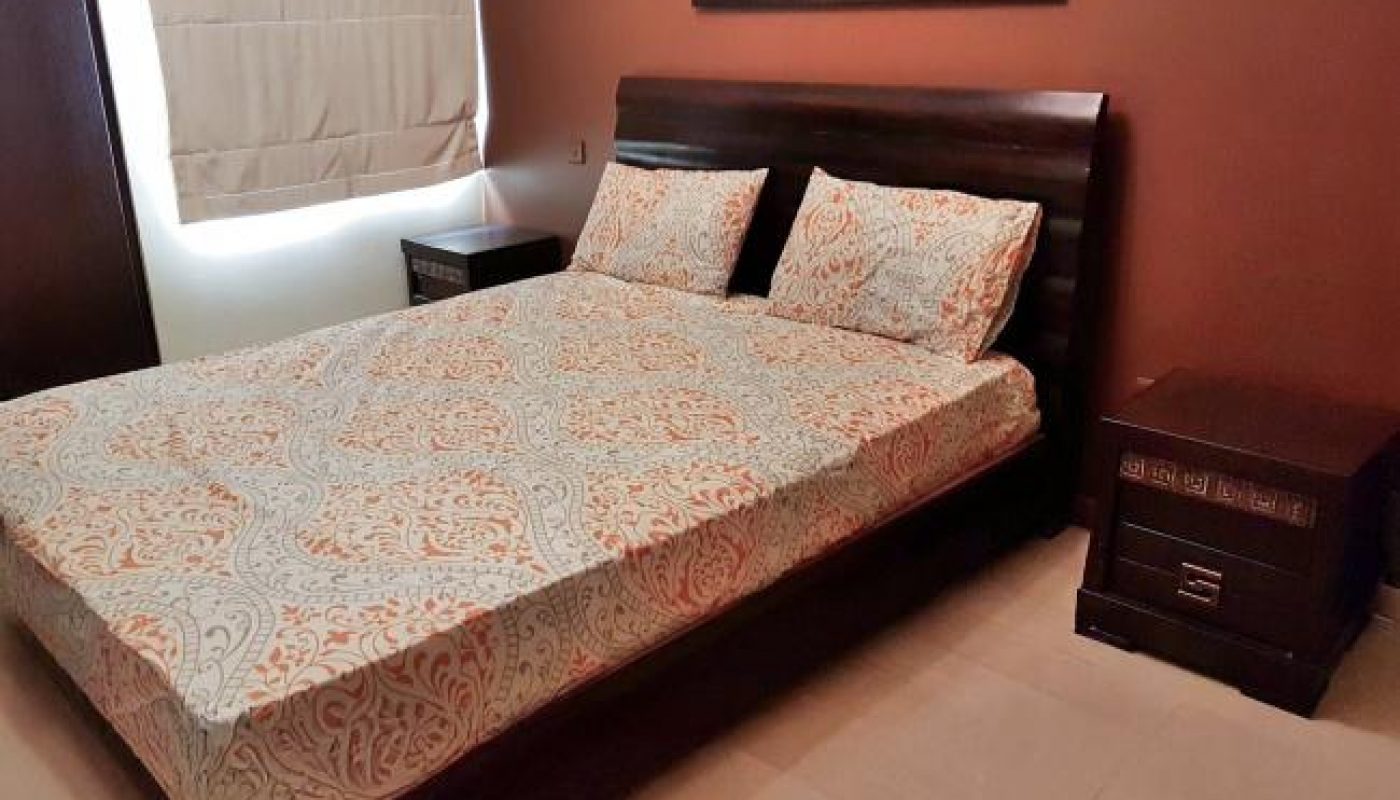 A neatly made bed with patterned bedding sits between two dark wooden nightstands in a bedroom with brown walls and a window covered by a beige shade.