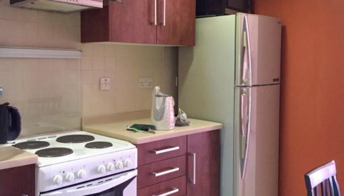 Modern kitchen with white stove, kettle, microwave, and stainless steel refrigerator, featuring wooden cabinets and an orange accent wall.