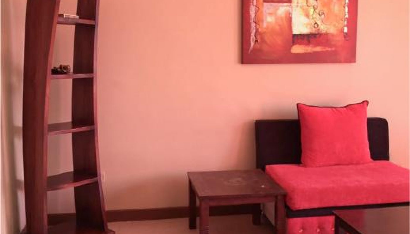 A small modern living room with a black and red chair, two wooden tables, a tall curved bookshelf, and an abstract painting on a peach-colored wall.