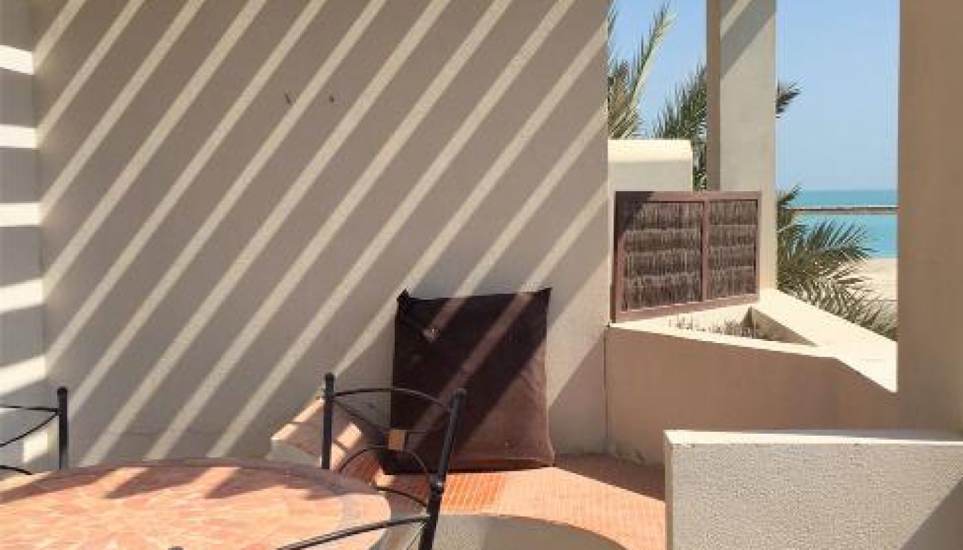 Outdoor patio with a round table and chairs under a wooden pergola, casting striped shadows; palm trees and sea visible in the background.
