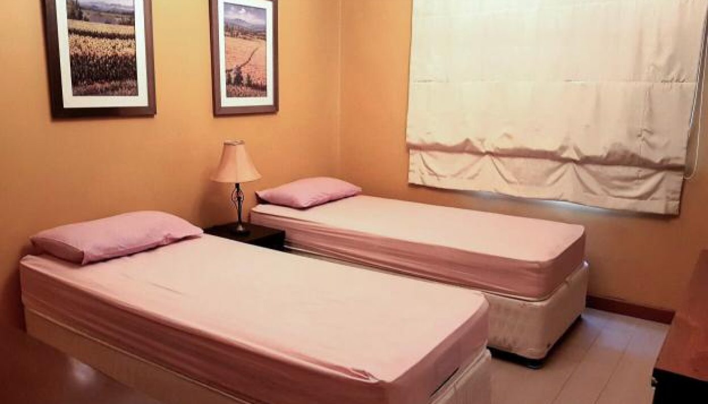 A small bedroom with two single beds, pink sheets, a lamp between them, two framed pictures on the wall, and a window covered by a white curtain.