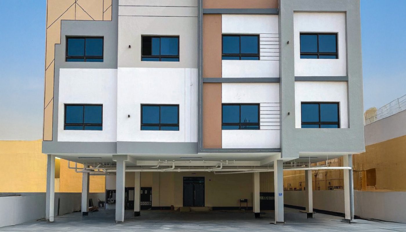 Three-story modern building with geometric design, large square windows, and an open ground floor supported by columns, set against a clear blue sky.