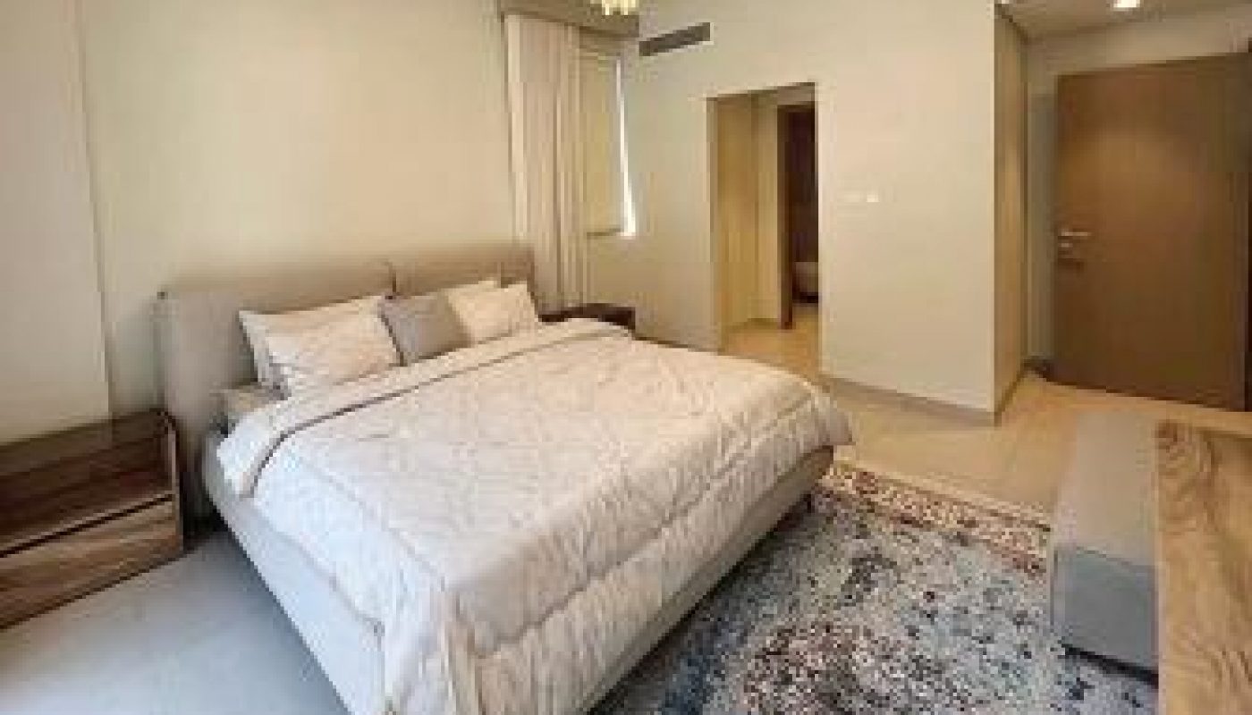 A neatly made bed with white bedding in a modern bedroom, featuring a chandelier, nightstands, a patterned rug, and light-colored walls.
