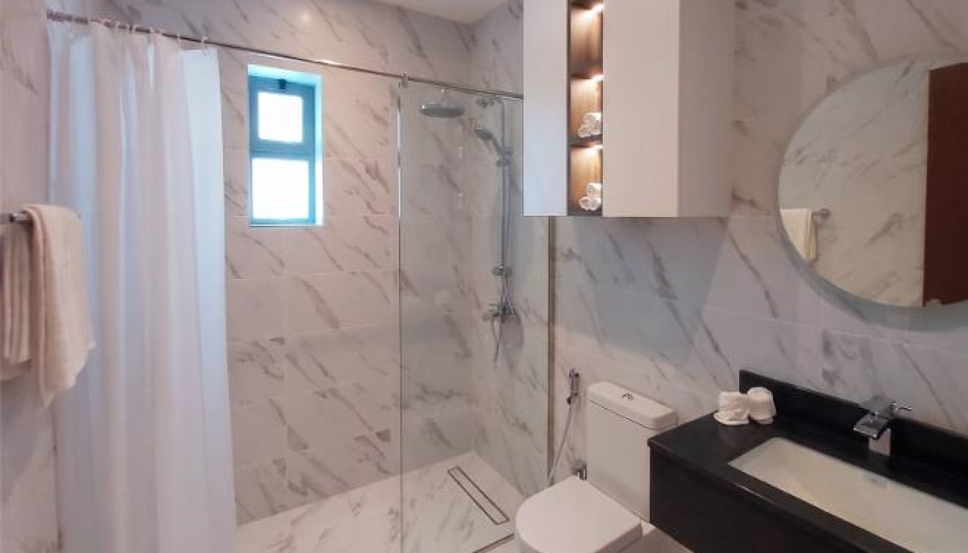 Modern bathroom with marble-patterned walls and floor, glass shower enclosure, wall-mounted sink, toilet, and storage cabinets, lit by ceiling lights and a small window.
