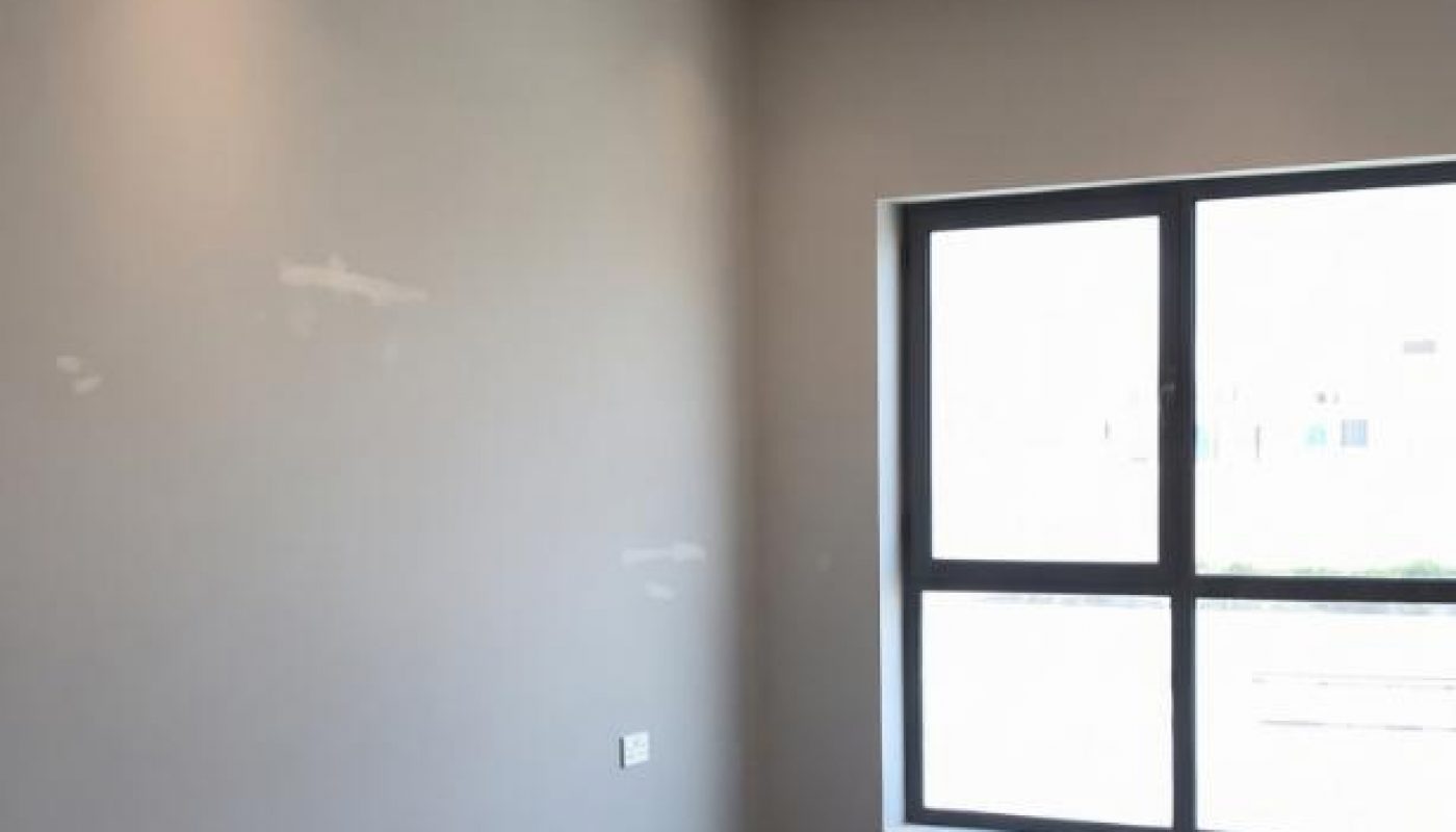 Empty modern room with light-colored walls, large window, recessed ceiling lights, and wood flooring.