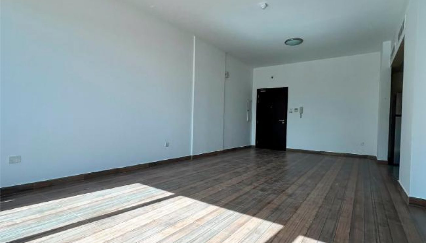 Empty room with white walls, wooden tile flooring, two ceiling lights, a black door, and sunlight streaming through unseen windows onto the floor.